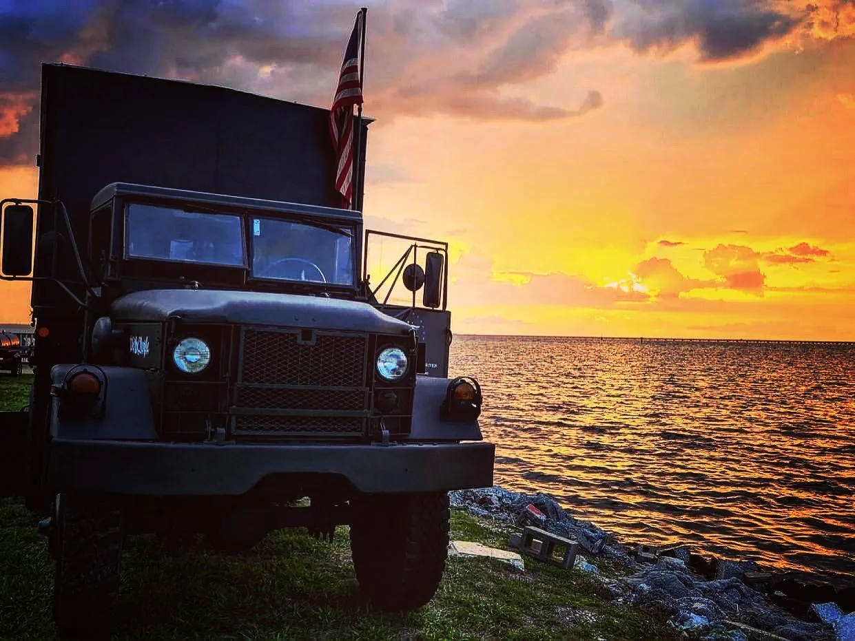 The Liberty Smokehouse truck, a 1971 military cargo truck flying the flag, parked by the water at sunset